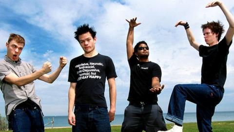 Sum41, St Kilda Beach Melbourne Australia. The four male members standing on green grass, wearing tshirt and jeans. They are making rock signs. The sky is cloudy blue.
