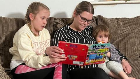 Woman in black and white top reads to 10-year-old and 2-year-old girls while they all sit on a brown sofa