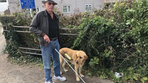 Roy Richards stood next to brambles with his guide dog and holding a cane