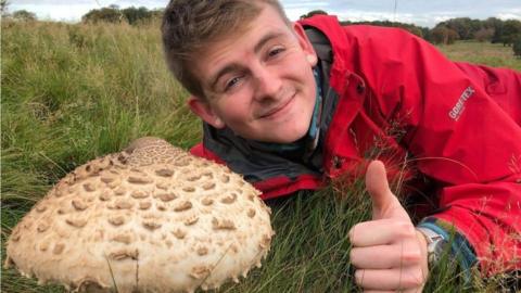 A man in a red coat gives a thumbs up while lying next to a large mushroom