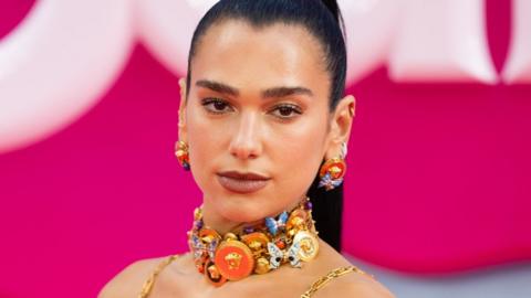 Dua Lipa at the London premier of the Barbie Movie in July 2023. The singer has her long hark hair pulled back into a high pony tail and wears a dark pink lipstick with neutral mkeup. She wears a large choker necklace made up of butterflies in gold, orange, blue and purple with matching earrings. She is photographed from the shoulders up in front of the pink Barbue logo and the gold straps of her dress can be seen over her shoulders.