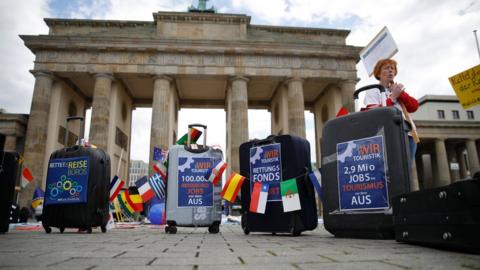Travel agency workers stand next to suitcases decorated with protest posters as they demonstrate on May 13, 2020 in front of Berlin's landmark the Brandenburg Gat
