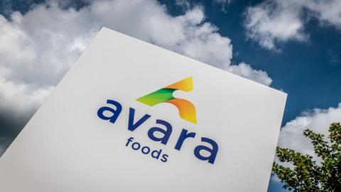 A picture of the Avara foods sign outside the Newent office, taken from below with blue sky and clouds in the background.