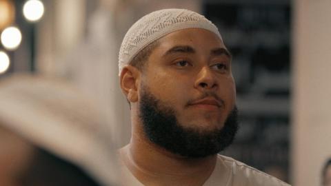 Big Zuu from from his his documentary Big Zuu Goes to Mecca. Big Zuu is a 28-year-old black man with shaved hair, a dark beard and brown eyes. He's pictured in traditional ihram clothing - white robes and a white skull cap.