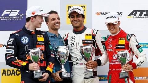 Enaam Ahmed is stood on a podium with three other racing drivers. He is standing second from the right wearing a white Mercedes racing driving overalls with a white cap. He is smiling and holding a silver trophy in his left hand. Stood next to him are three white racing drivers who are also all wearing racing overalls and holding silver trophies and smiling. Behind them is a sponsors board with various racing sponsors on it.