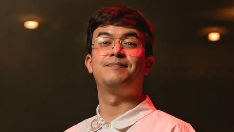 A man with short black hair and glasses stands on a stage as a darkened theatre auditorium stretches out behind him. He's wearing a white shirt and looks relaxed.