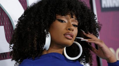 Megan Thee Stallion attends the CMT (Country Music Television) Music Awards in Austin, Texas, U.S., April 2, 2023. Megan, a black woman in her late 20s, has shoulder length afro hair and holds her manicured hand in it. She wears large white hoop earrings and a blue high necked top. She has winged black eye liner and and glittery eye shadow and pink lipgloss.