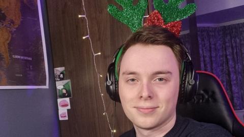 A man sits facing the camera while smiling wearing headphones and festive sparkly green reindeer antlers