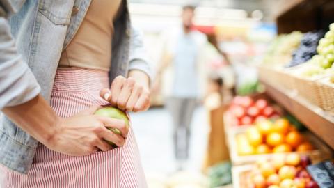 Close-up of unrecognizable woman in stripped skirt hiding apple in pocket while stealing it in food store