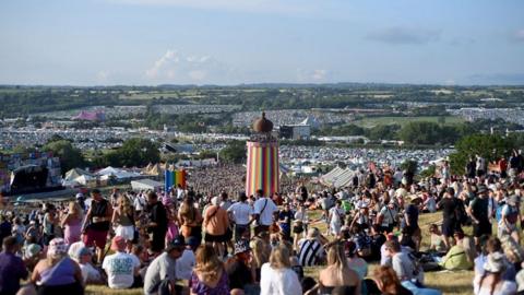 Festival goers sit on the hill at Glastonbury over-looking fields full of people, tents and a tall striped tower in the middle. The sky over the horizon is a grey blue colour