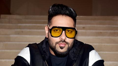 Badshah looking at the camera wearing a black leather jacket with white shoulder pattern. Hee's wearing yellow tinted glasses with his hair slicked up. The background is of light brown coloured long steps.