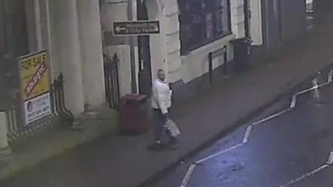A man wearing a light-coloured jacket holding a shopping bag walking on the pavement on Bedminster Parade