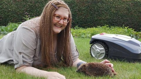 Dr Sophie Lund Rasmussen laying on the grass with a hedgehog next to a robot mower