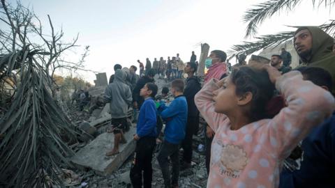 Palestinians watch as people search for bodies and survivors among the rubble following Israeli airstrikes on the west of Al Nusairat refugee camp, southern Gaza Strip, 20 February 2024
