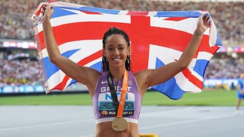 Katarina Johnson-Thompson stood at the side of an athletics track holding a Union Jack flag above her head and behind her with both arms. She is smiling and has her black hair in a ponytail behind her back. She is wearing a purple and white cropped Team GB vest and has a medal around her neck.