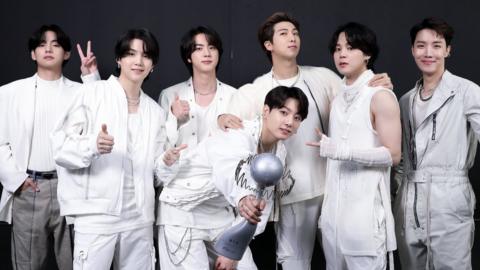 All seven members of BTS are posing for a photo, having won an award. The Korean boyband are dressed in white, posing with smiling faces, with a black background behind all seven of them