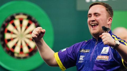 A darts player celebrates, with a dart board in the background. He wears a purlpe and yellow short-sleeved T-shirt, with three darts in his lift hand.