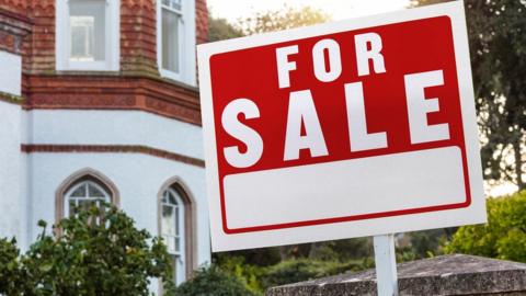 A red sign with white writing saying "for sale" stands outside a townhouse