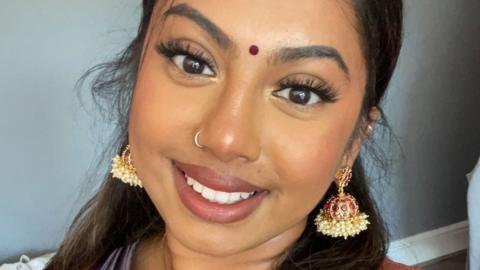 Kirthana - a 21-year-old south Asian woman, smiles at the camera as she takes a selfie. She has her long, dark hair styled half-up-half-down and wears a bindi and her makeup includes a glittery gold eyeshadow and false eyelashes. She wears decorative dangly earrings which comprise a round red ornament decorated in gold and pearls. She is pictured inside against a pale blue wall.