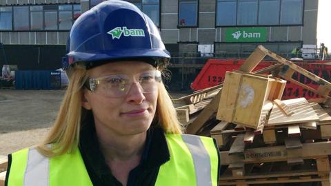 A woman wearing a protective hard hat and safety goggles with a building and a pile of wood behind her