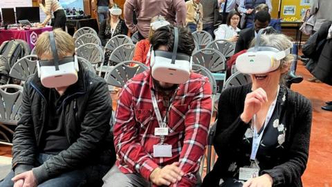 Three people wearing white VR headsets. The one on the right is laughing and appears to be raising her hand to touch something that is not there.