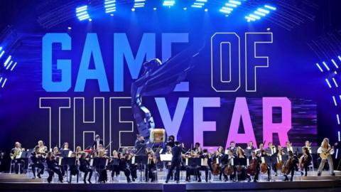 An orchestra performs on a large stage with "Game of the Year" projected on to a screen at the rear in colourful block capital letters. An image of the awards statue - a winged figure leaning backwards as if about to take flight - is superimposed on top. There are between 30 and 40 members of the orchestra, most of them seated and playing a range of stringed and woodwind instruments. To the far right, an electric guitarist stands facing the crowd, and a large cylindrical Japanese Taiko drum is on a plinth in the centre of the group.
