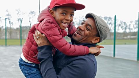 In a playground, a dad with a flat cap lifts his young son with a baseball cap, both smiling.