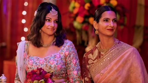Characters of Kamala and Nalini standing next to each other looking slightly to their left, dressed in wedding attire. Kamala is wearing a shiny floral top, while holding flowers in her hand, with a jewellery item in her hair going down to her forehead. Nalini is wearing a pink shiny outfit, with earrings. The background is red decorated with flowers.