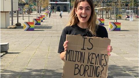 Anouska Lewis holds a sign saying "Is Milton Keynes Boring?"