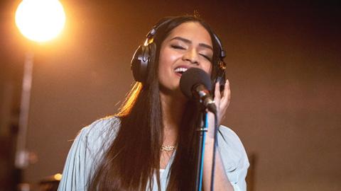 Saloni, an Asian woman, closing her eyes and singing into a black microphone while wearing black headphones. Behind her is black background and a spotlight shining on her. The top part of her outfit is light blue.