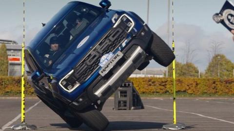 A man with a clipboard watches as a pickup on two wheels is driven between