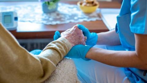 A carer in surgical gloves holds an old woman's hand