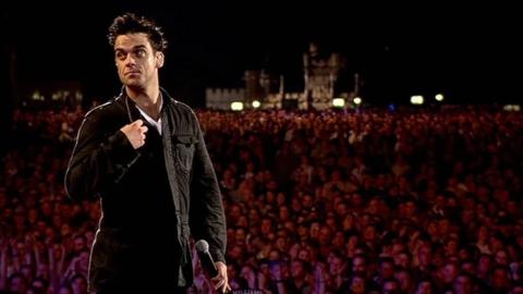 Robbie Williams performing at Knebworth Park in 2003. He is facing the back of the stage with a huge crowd behind him.
