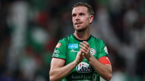 Jordan Henderson reacts after the Saudi Pro League match between Al-Ettifaq and Al-Ittihad at Al Ettifaq Stadium on November 24, 2023 in Al Dammam, Saudi Arabia. Henderson is a 33-year-old white man with short brown hair and blue eyes. He wears a green Al-Ettifaq kit and a red arm band. He is pictured clapping on the pitch with a crowd blurred out behind him.