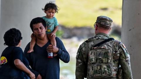 A man carries his daughter on his shoulders as a group of migrants are apprehended by US Border Patrol and National Guard troops in Eagle Pass, Texas, near the border with Mexico on June 30, 2022