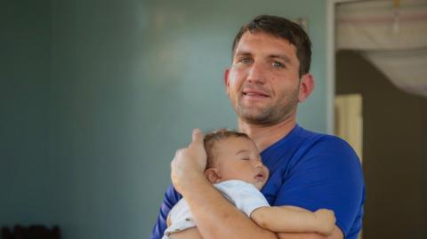 Rafael cradling his baby outside his tiny house