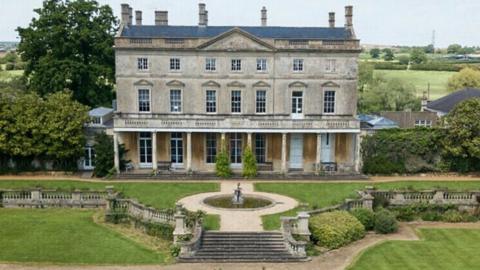 A large period building with stone pillars, surrounded by gardens and hedgerows