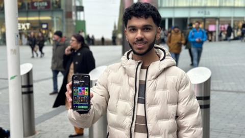 A man wearing a beige puffa jacket stands in front of a shopping centre holding a phone showing a screen with the Roblox app on it