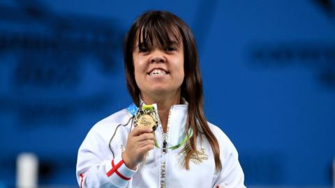 Zoe Newson in an white England branded zip up top. Ms Newson is holding a Gold commonwealth medal and smiling.
