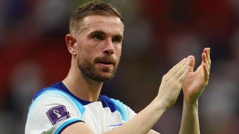 Jordan Henderson celebrates for England by clapping his hands. He is wearing a white England jersey with blue pattern going around the neck and shoulder line, with a World Cup logo on his right arm.