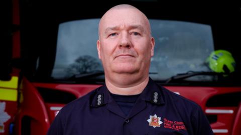 Terry Maher in front of Colchester fire station with a fire engine behind him