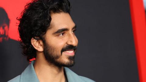 Dev Patel on a red carpet for a Monkey Man premiere. Dev is a 33-year-old British Asian man with curly dark hair, brown eyes and a short beard and moustache. He wears a pale blue suit as he poses for photos on the red carpet in front of red and black hoarding advertising his new film