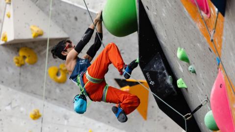 Anita Aggarwal climbing up a wall and hanging on to a green boulder
