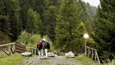 Two young men stand on a mountain gravel track on the Italian side of the French-Italian border, wearing trainers and carrying a small bag
