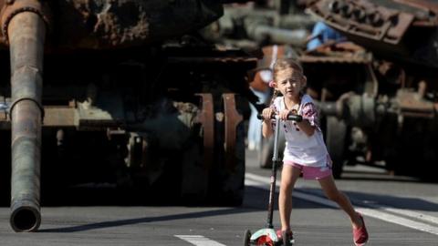 Girl on scooter in front of armoured tanks