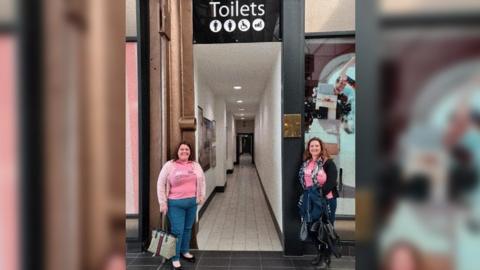 Louise and Jessica from Mums On A Mission at the MacArthur Glen designer outlet in Rodbourne, Swindon. They are stood at the entrance of a corridor leading to the toilets. A black sign reading "toilets" is above their head, and there are symbols below the text, showing a male, female, baby changing and disabled toilet. Both women have long dark hair and are wearing blue jeans and a pink t-shirt. They are smiling and looking at the camera.