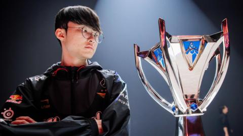 A young Korean man with black hair and large round glasses stands next to an elaborate trophy under a stage spotlight. His arms are folded and he has a serious look on his face. He wears a black tracksuit jacket with various sponsor logos - such as Red Bull - on the arms. The silver trophy has a hourglass shape - wide at the bottom, narrow in the middle and wide at the top, with four “arms” acting as handles extending out from the centre to the top. A blue gem is placed at the point where each arm joins.