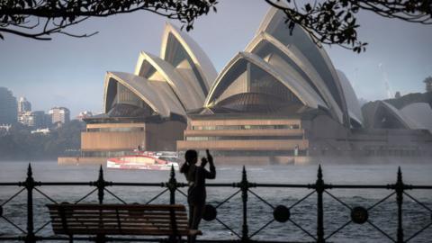 A woman takes a picture of the Sydney Opera House and Sydney Harbour