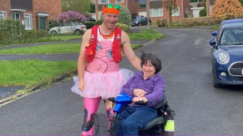 Phil Wiseman on the street wearing a pink tutu and leggings, next to his mum who is in a motorised wheelchair