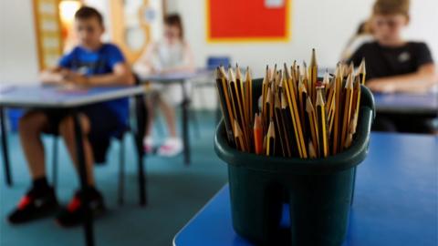 A pot of pencils in a classroom, with children sitting at desks in the background
